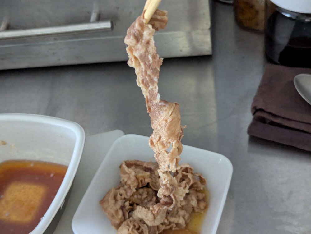 吉野家の牛丼の具から、箸で大きな牛肉スライス1枚を持ち上げている場面。白い器には盛り付け済みの牛丼の具、奥には残った汁の入った容器が見える。