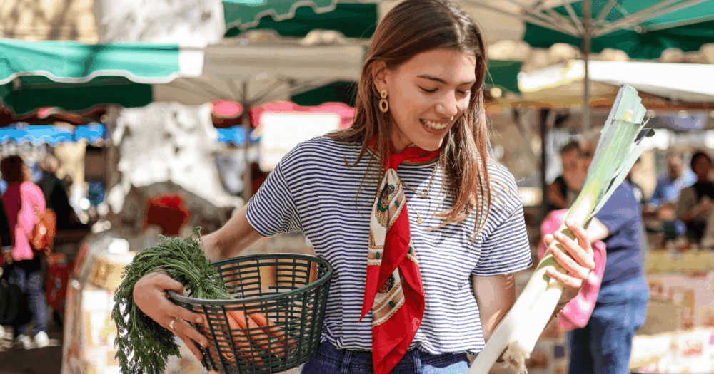 市場でカゴを持ち、ニンジンと長ネギを手に笑顔で野菜を選ぶ女性